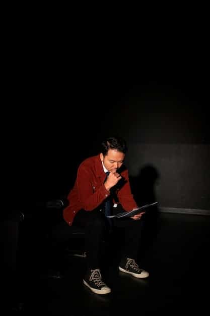 A film critic sitting in a dark movie theater, illuminated by the glow of a laptop screen as they type notes. The screen shows a paused scene from a recent drama film.